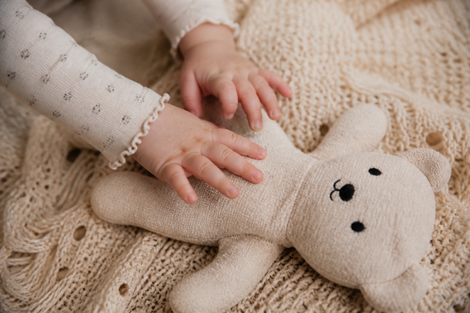 Kleine Babyhände greifen nach einem hellen, flachen Teddybären mit gestickten Augen auf einer Krabbeldecke