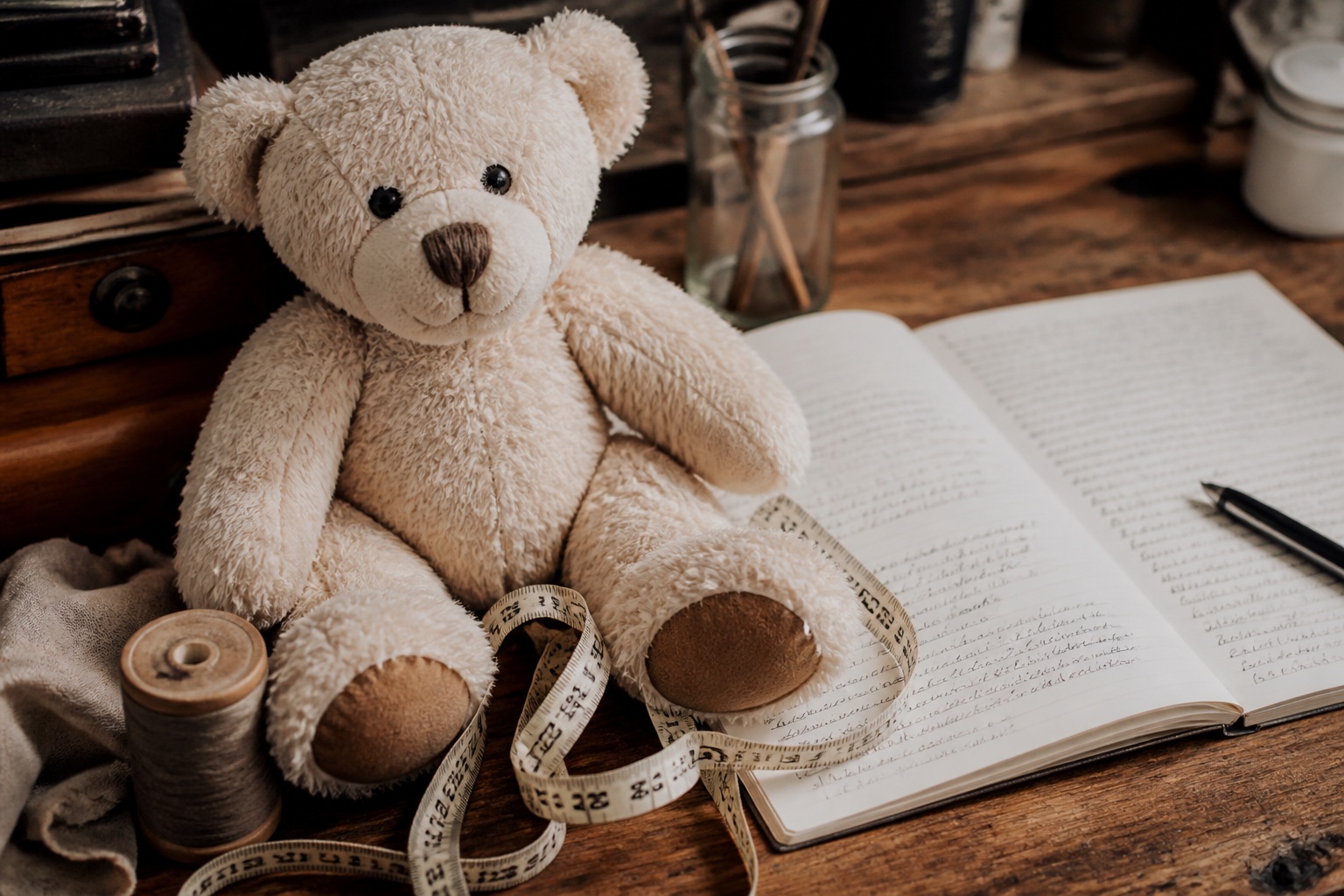 Schreibtisch mit einem hellen Teddybären, einem aufgeschlagenen Notizbuch, einem Maßband und einer Garnrolle